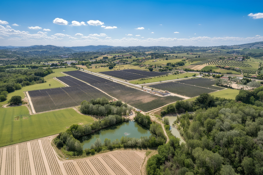 High-angle aerial view of the Oasi Barberini solar panel fields and a natural pond, surrounded by hills and trees.