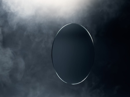 Clear oval optical lens remaining perfectly transparent while surrounded by dense white smoke and fog in a dark studio.